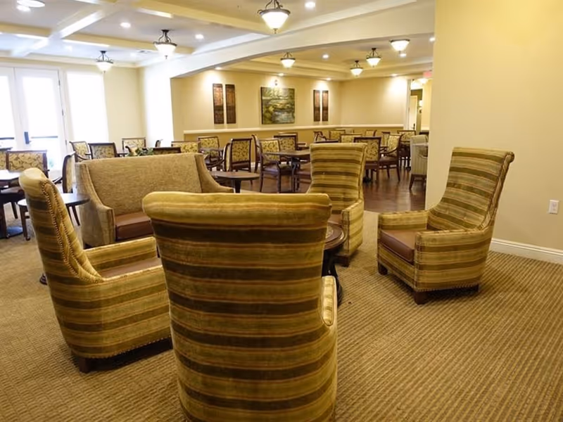 A cozy seating area in a senior living facility with four striped armchairs arranged around a small round table, a loveseat, and several dining tables and chairs in the background. The room is warmly lit with ceiling lights and decorated with framed artwork on the walls.