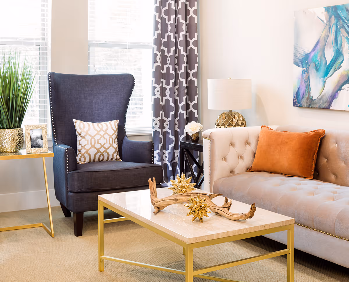 A bright living room with a dark blue armchair featuring a patterned cushion, a beige tufted sofa with an orange pillow, a gold and marble coffee table with decorative items, a side table with a gold plant pot and framed photo, a lamp on a black side table, patterned curtains, and abstract wall art.