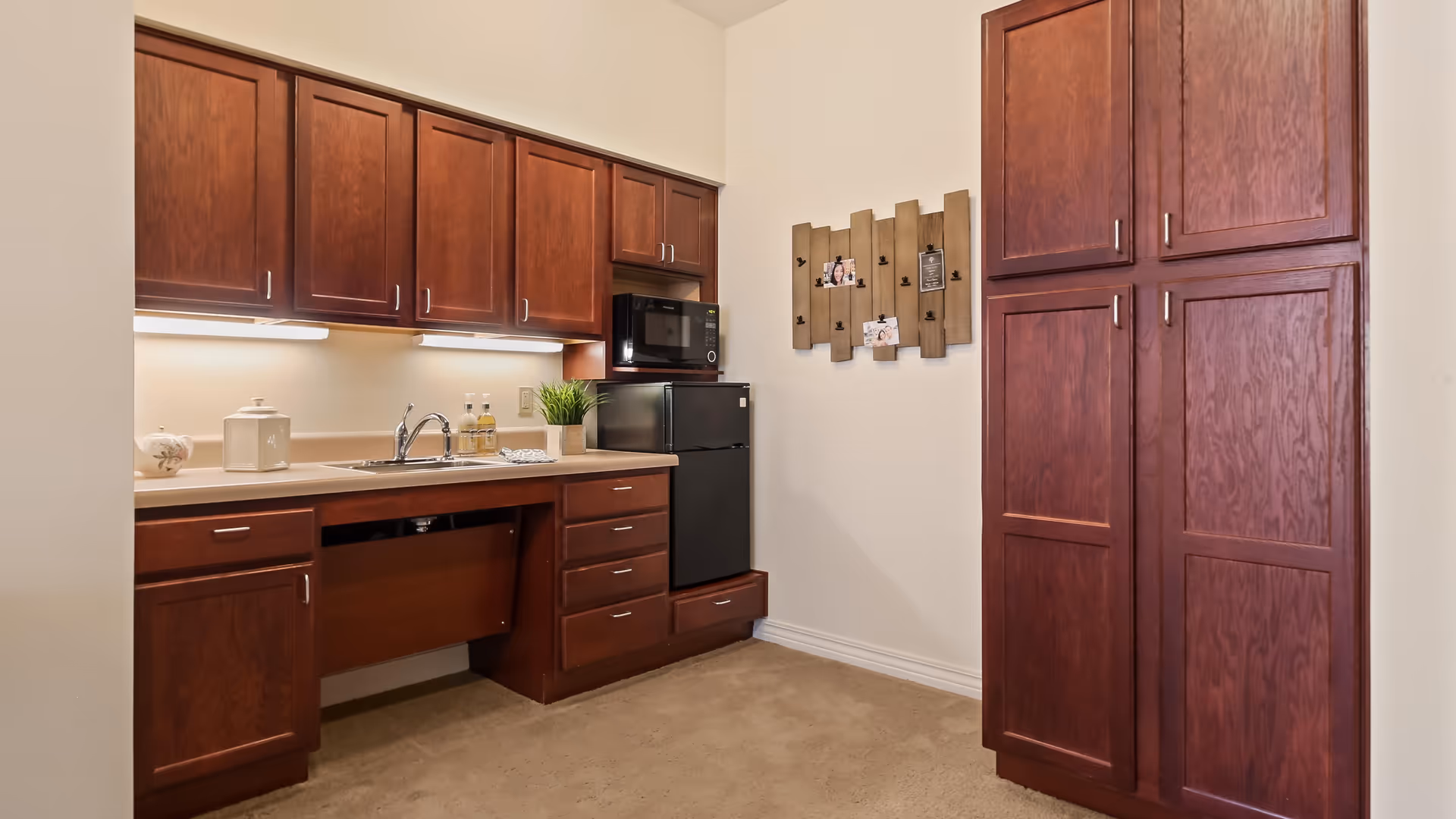 A compact kitchen area with dark wood cabinets, a small black refrigerator, a microwave, a sink with a faucet, and under-cabinet lighting. There is a decorative wooden wall hanging with photos on the adjacent wall.