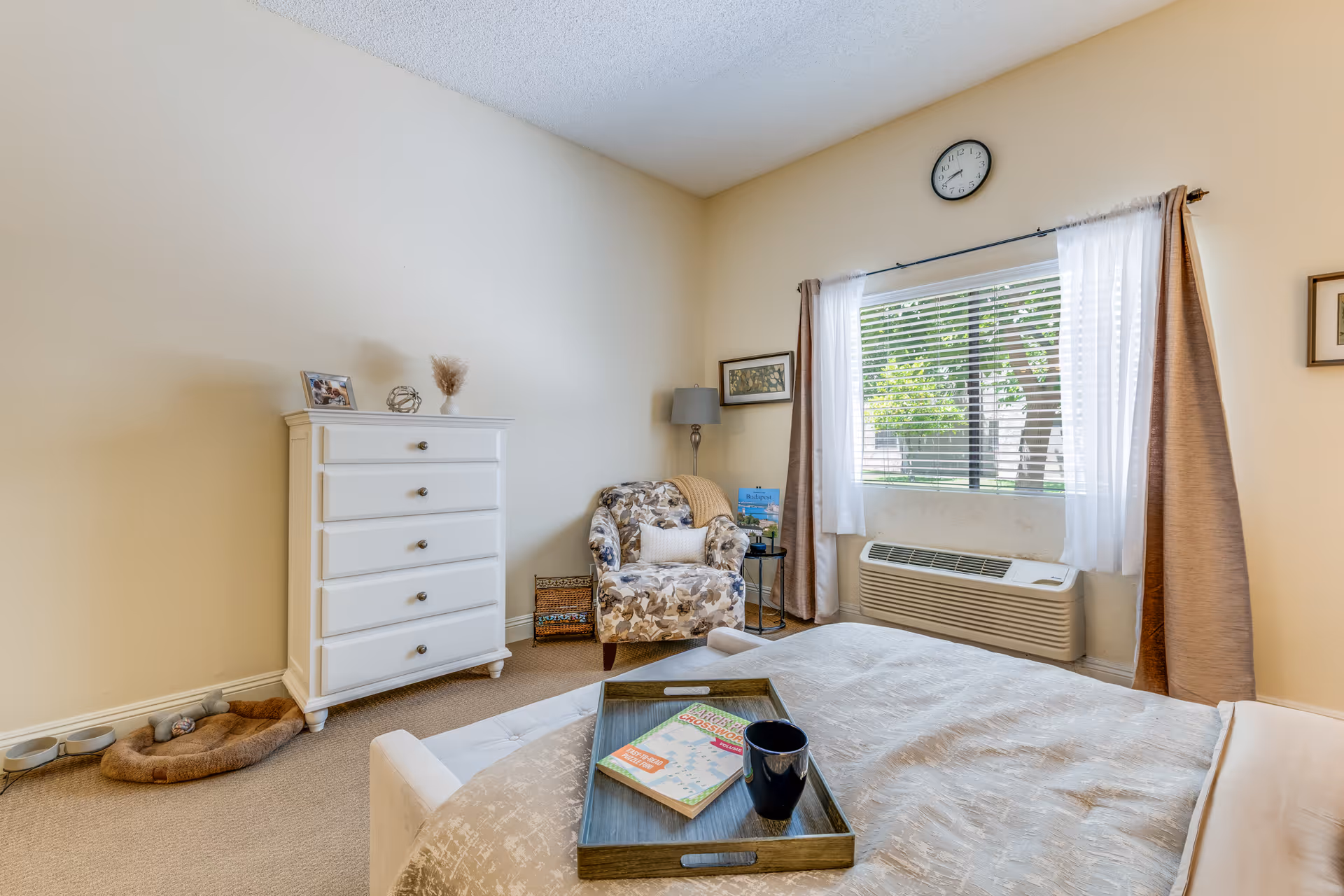 A cozy bedroom with a bed covered in a beige comforter, a wooden tray with a book and a mug on the bed, a floral armchair next to a small round table with a lamp, a white chest of drawers with decorative items on top, a window with white and brown curtains, and a wall clock above the window.