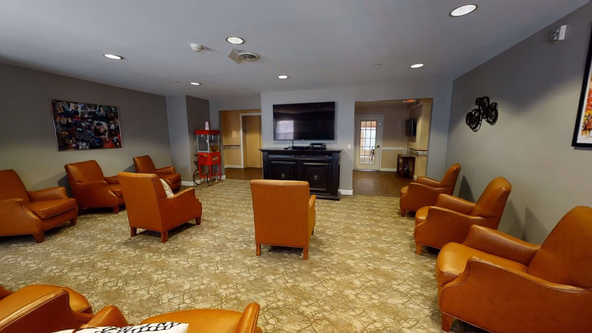 A spacious living room with multiple brown leather armchairs arranged in a semi-circle facing a large flat-screen TV mounted on a white wall above a dark wooden cabinet. The room has beige patterned carpet, gray walls, recessed ceiling lights, and decorative wall art. A red popcorn machine is visible in the corner near a hallway entrance.