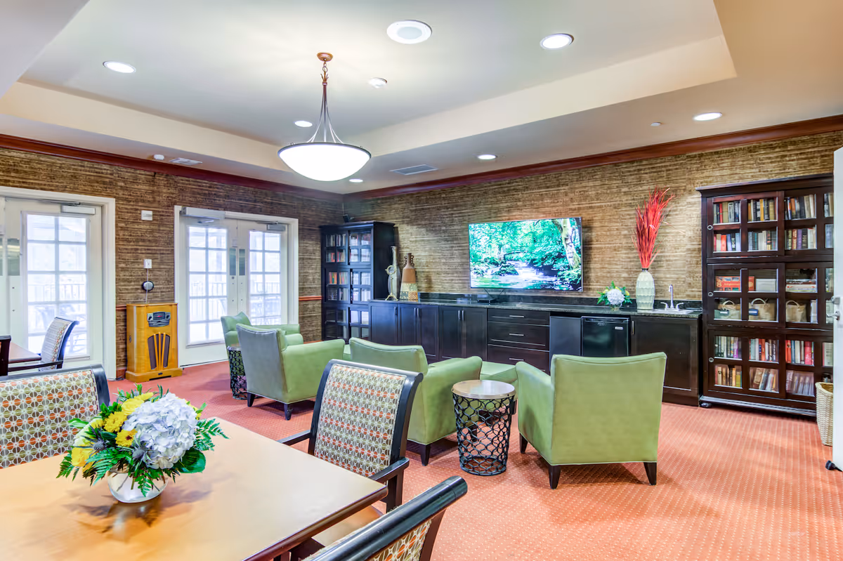 A cozy common area in Arbor Terrace Crabapple featuring a seating arrangement with four green armchairs around a small round table, a large flat-screen TV mounted on a wall with dark cabinetry below, two tall bookshelves filled with books and decorative items, a wooden table with patterned chairs and a floral centerpiece, and double glass doors letting in natural light.