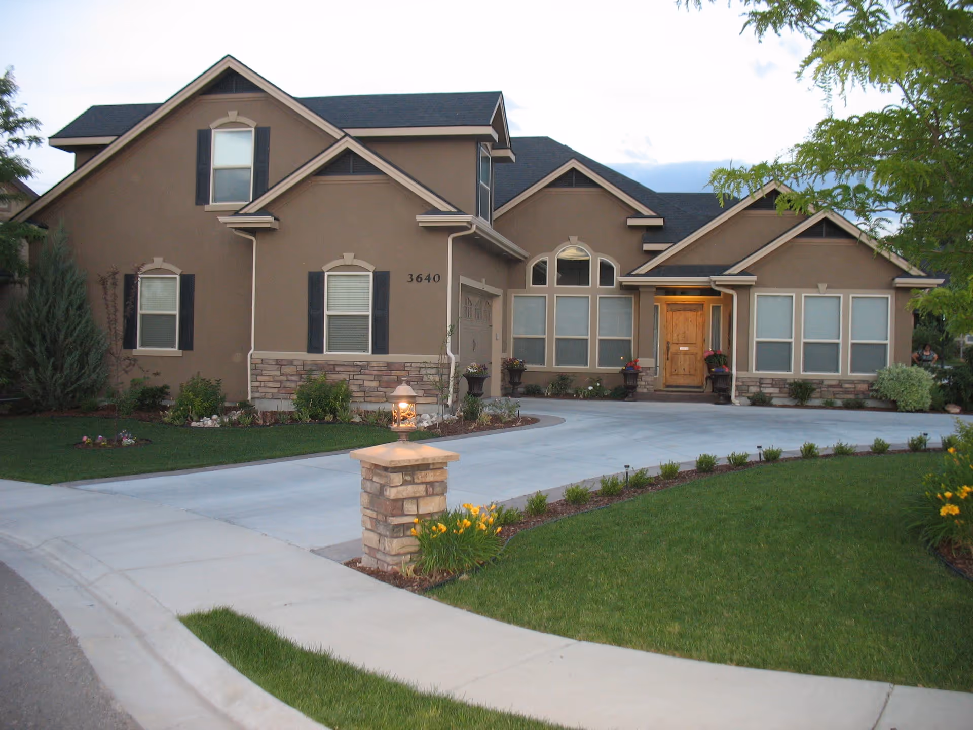 Front exterior view of a single-story residential building with a beige facade, multiple windows with white blinds, a wooden front door, and a well-maintained lawn with flower beds and a stone lamp post near the driveway.