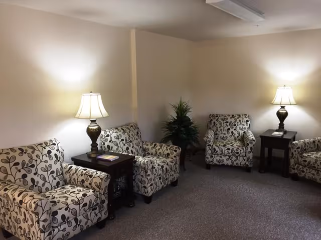 A cozy sitting area with four patterned armchairs arranged around two wooden side tables, each with a lamp on top. There is a green potted plant in the corner, and the room has beige walls and carpeted flooring.