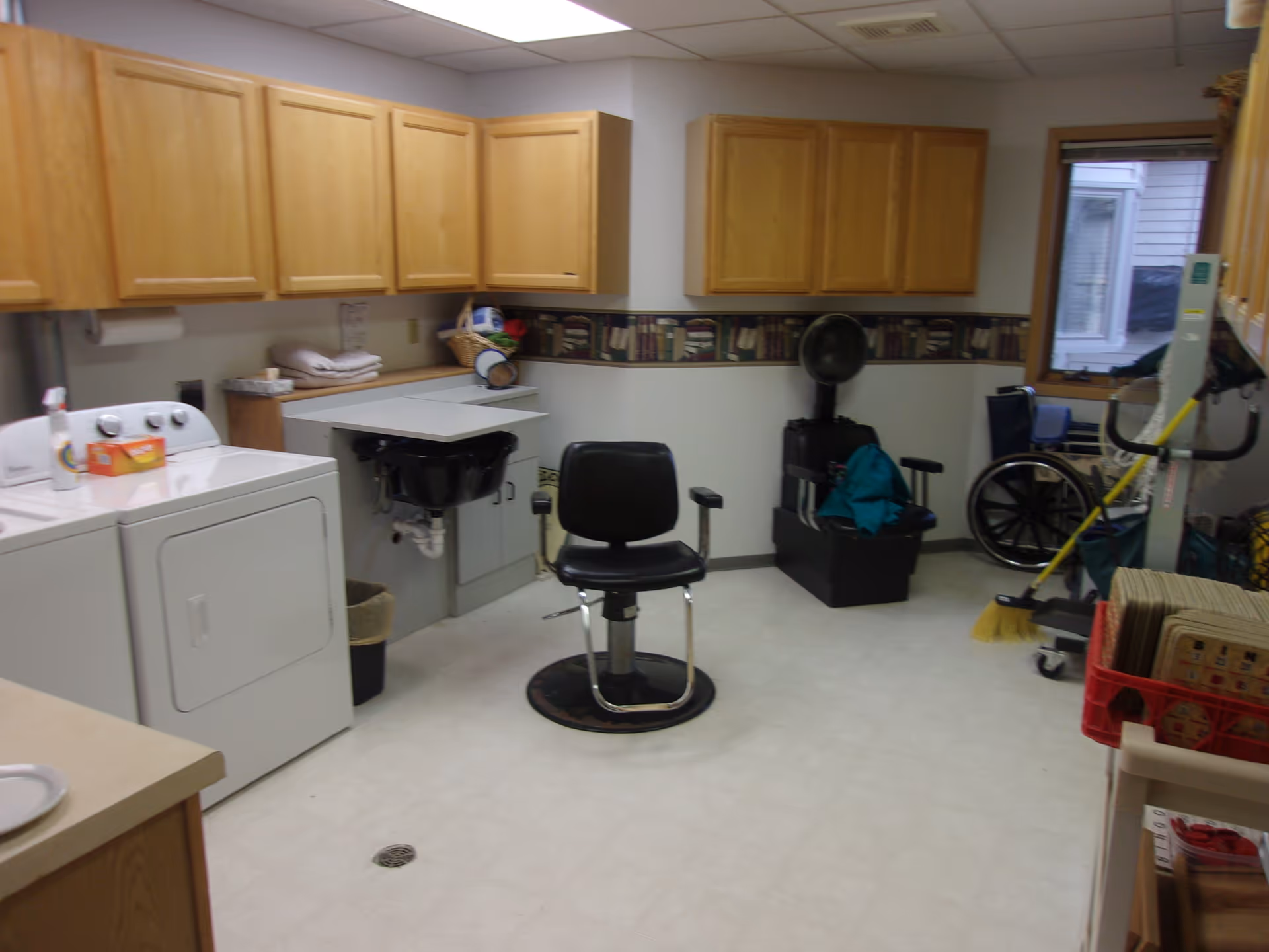 A multipurpose room with wooden cabinets mounted on the walls, a washing machine and dryer, a black salon chair, a hair washing station, a hair dryer chair, a wheelchair, cleaning supplies including a broom, and various other items. The room has a window and a tiled floor.