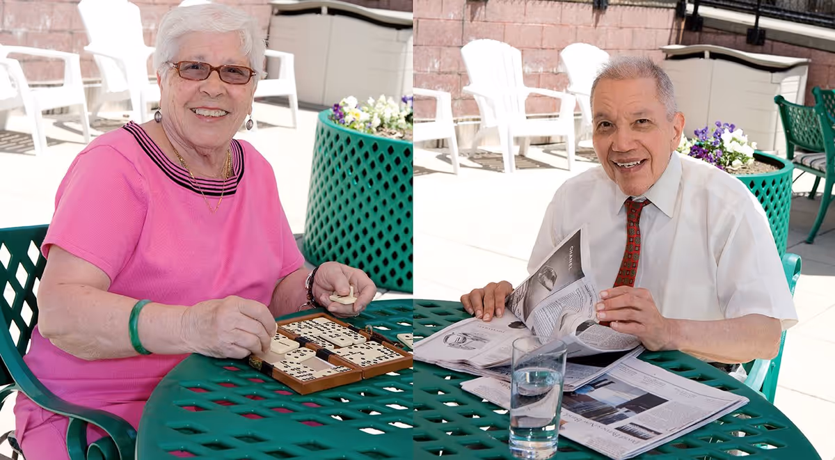 An elderly woman and an elderly man sitting at a green outdoor table. The woman is smiling and playing dominoes, while the man is smiling and reading a newspaper. They are seated on green chairs with white plastic chairs and flower pots visible in the background on a sunny patio.