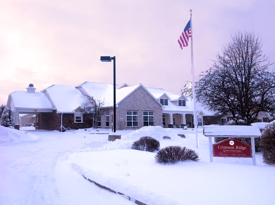 Exterior view of Crimson Ridge Meadows senior living community building covered in snow with an American flag on a flagpole and a sign displaying the community name in front.