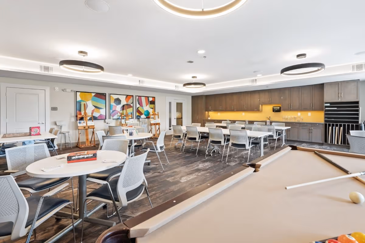 A spacious, modern common area with multiple tables and chairs arranged for group activities or dining. The room features a pool table in the foreground, colorful abstract artwork on the walls, and a long counter with cabinets and a yellow backsplash in the background. The ceiling has circular light fixtures, and the floor is dark wood.