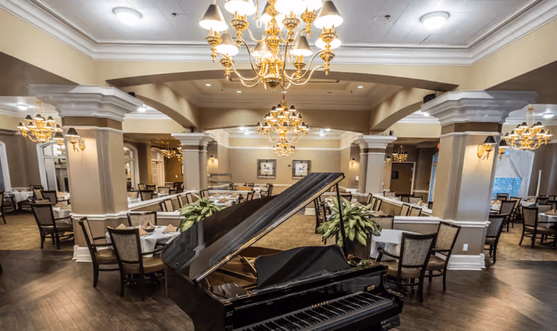 Elegant dining room with a grand piano, chandeliers, and multiple tables set for meals.
