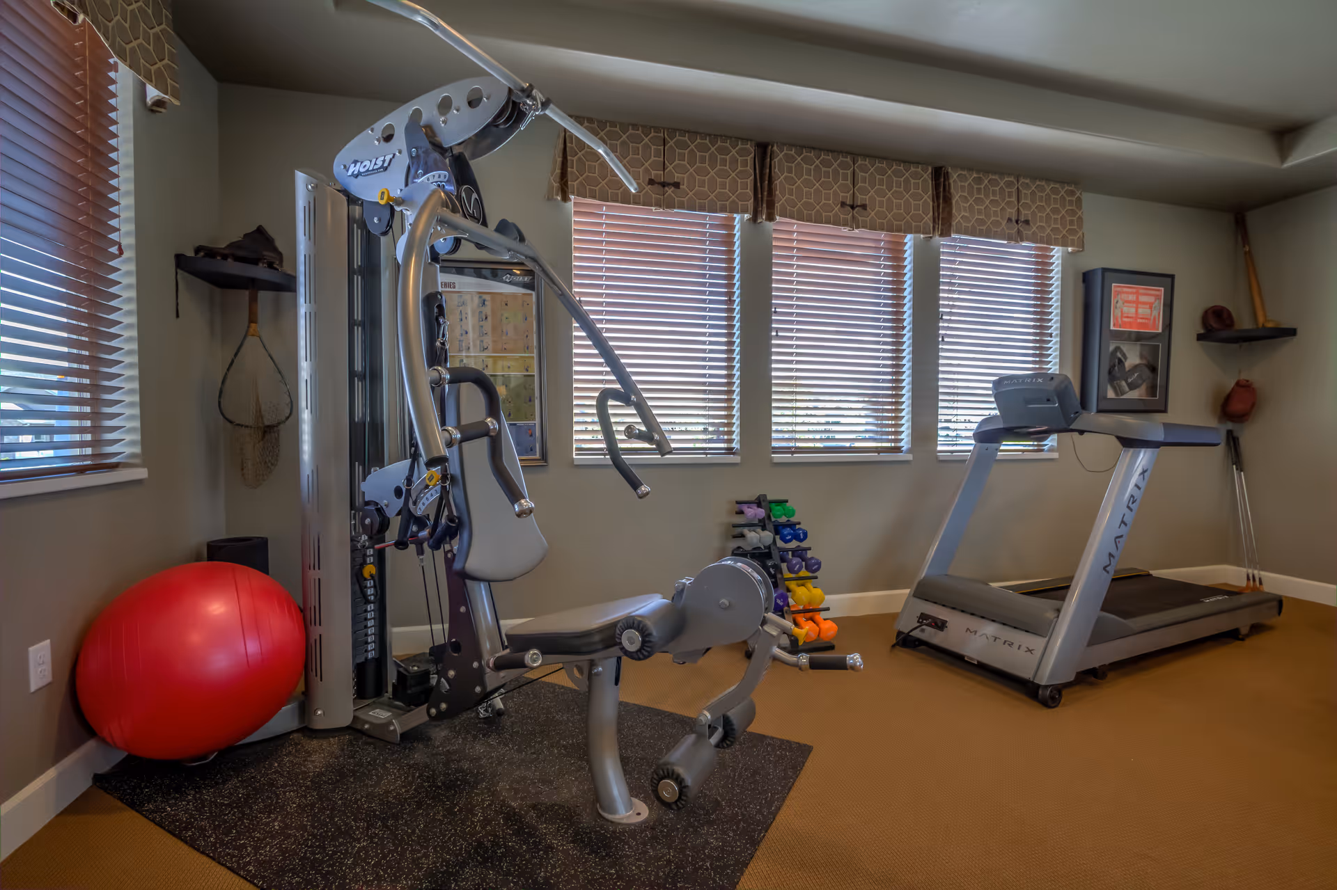 A fitness room with exercise equipment including a multi-function weight machine, a treadmill, a rack of colorful dumbbells, and a large red exercise ball. The room has beige walls, carpeted floor, and windows with wooden blinds letting in natural light.