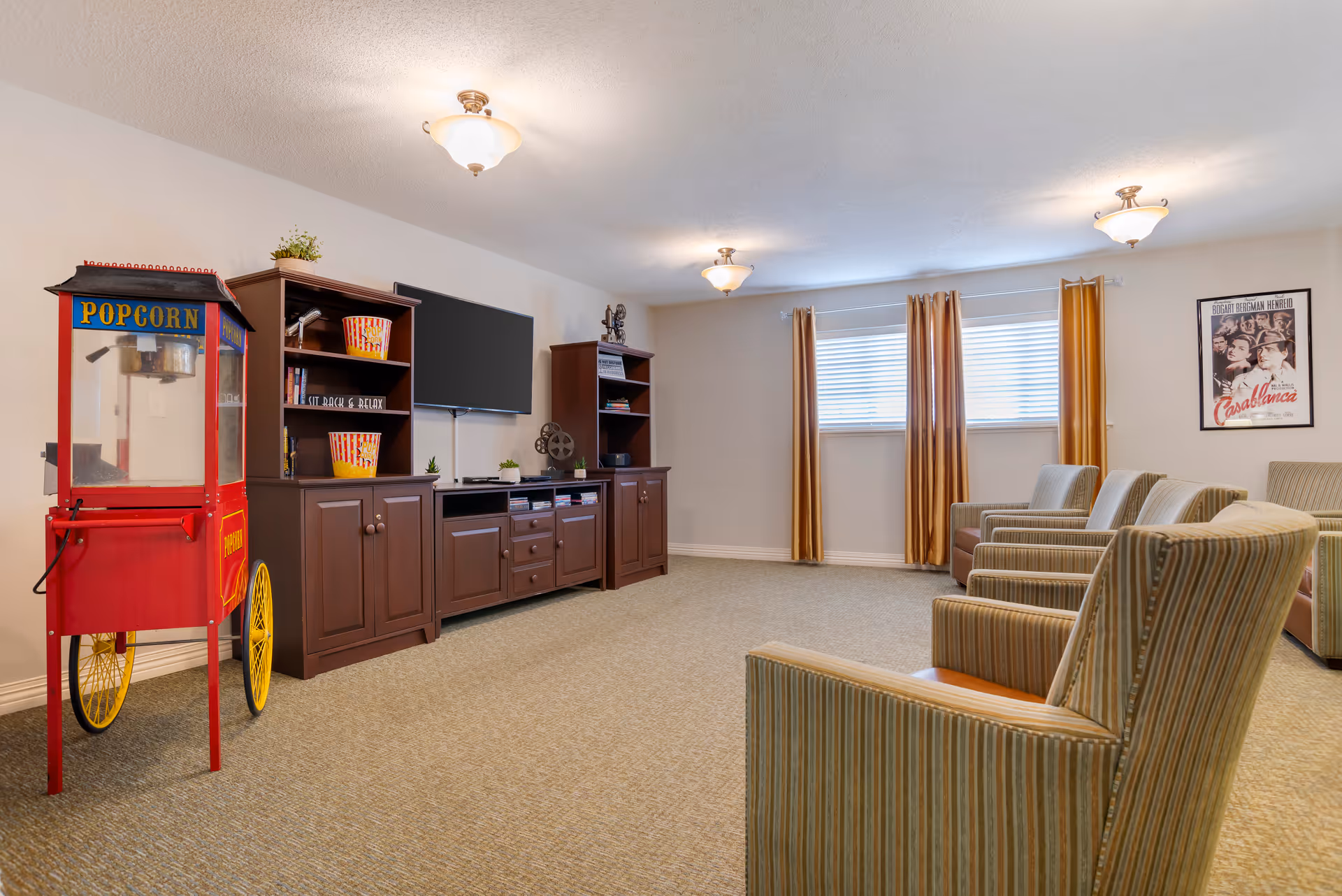 A cozy entertainment room with a vintage-style red popcorn machine on the left, a dark wood entertainment center with a flat-screen TV mounted above it, and shelves holding popcorn buckets and decorative items. There are several striped upholstered armchairs arranged facing the TV. The room has beige carpet, cream-colored walls, three ceiling lights, a window with gold curtains, and a framed Casablanca movie poster on the wall.