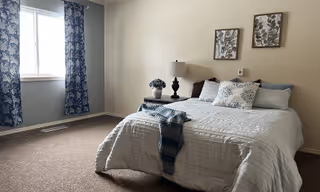 A neatly made bed with white and blue bedding in a simple bedroom. The bed has multiple pillows and a blue throw blanket. There is a bedside table with a lamp and a small decorative item. Two framed floral artworks hang on the beige wall above the bed. A window with blue patterned curtains lets in natural light.
