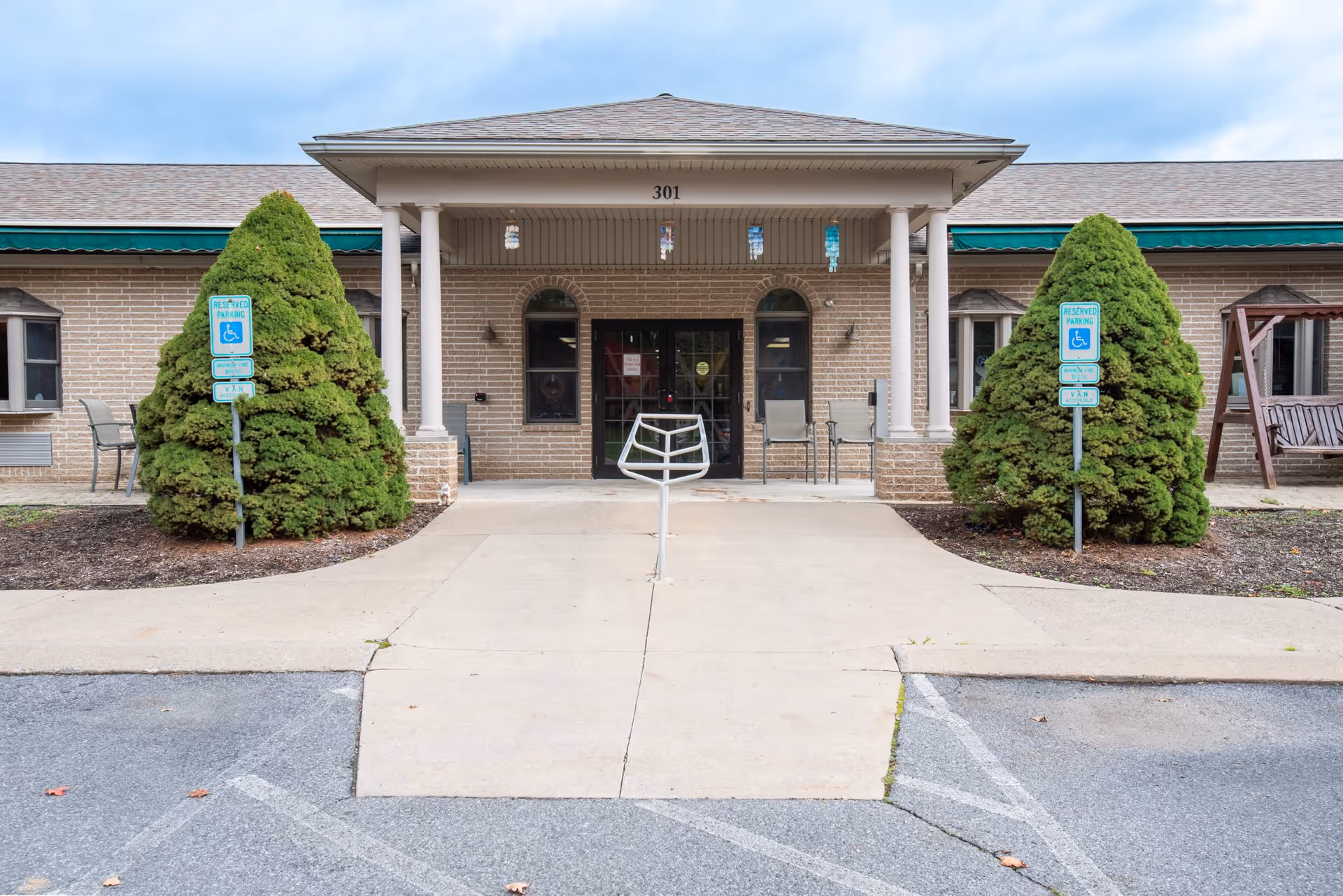 Front entrance of a single-story brick building with the number 301 above the door. There are two large green bushes on either side of the entrance, each with a reserved parking sign for handicapped access. The entrance has a covered porch supported by four white columns, and there are chairs placed near the door. The sky is partly cloudy.