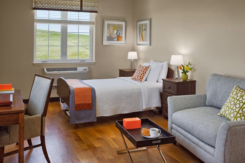 A cozy bedroom in a senior living facility featuring a single bed with white bedding and an orange throw, two bedside tables with lamps, a gray upholstered armchair with a green patterned pillow, a wooden desk with a chair, and a window with a view of green grass outside.
