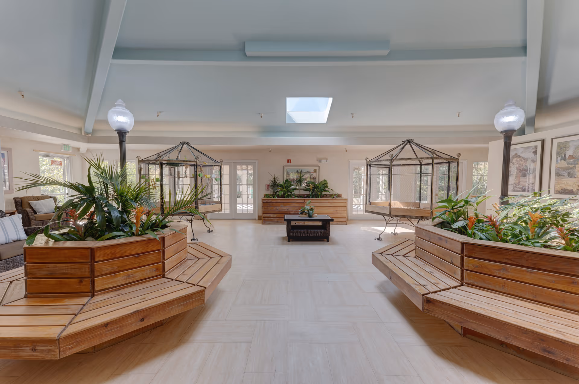 A spacious indoor common area with wooden bench seating surrounding large planters filled with green plants. Two hanging wicker chairs are suspended from metal frames in the background. The room has a high ceiling with a skylight and is well-lit with natural light coming through multiple glass doors and windows. There are also two vintage-style lamp posts inside the room and framed artwork on the walls.