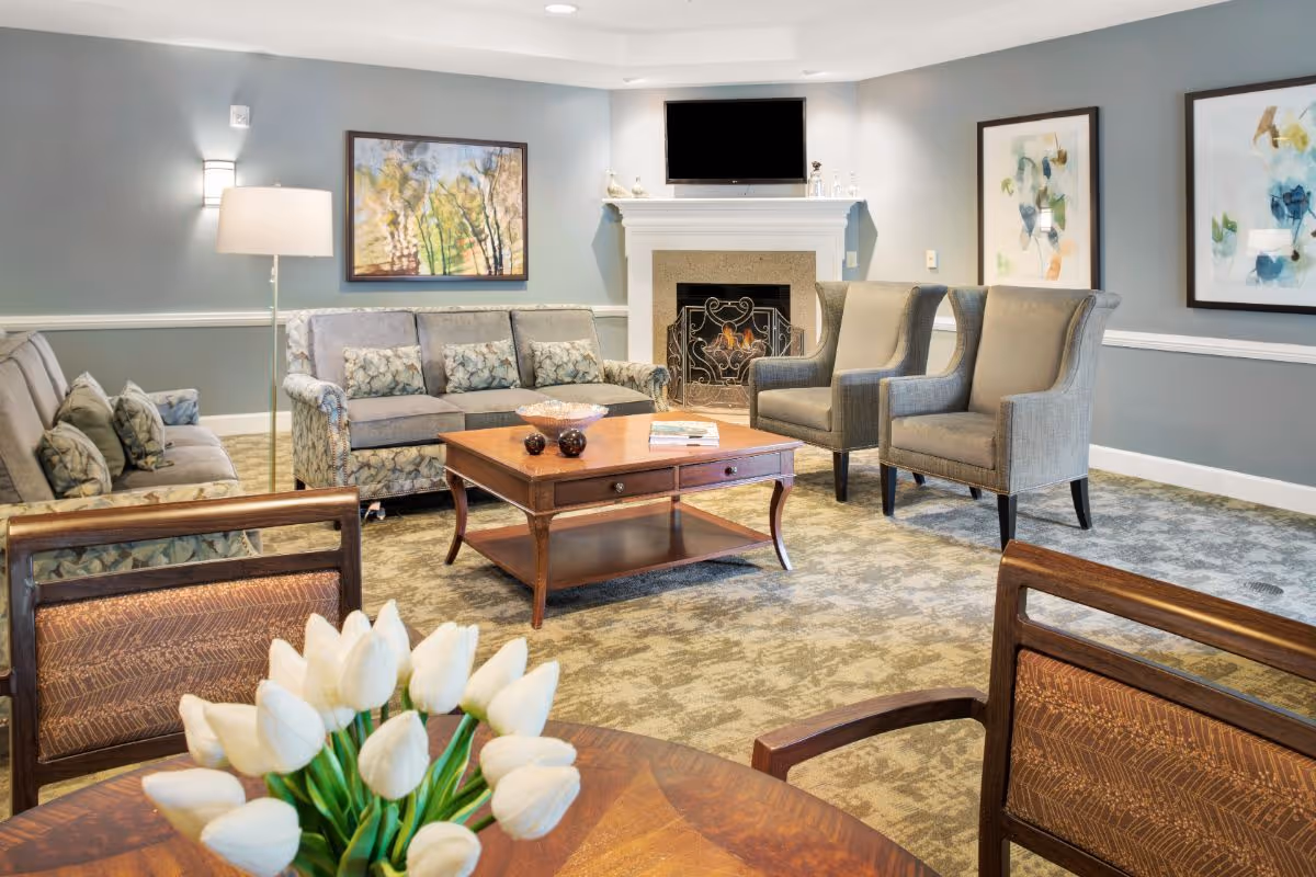 A cozy living room area with two gray sofas, two gray armchairs, a wooden coffee table, and a fireplace with a TV mounted above it. There are framed abstract paintings on the walls and a floor lamp beside one sofa. In the foreground, there is a wooden table with a bouquet of white tulips.