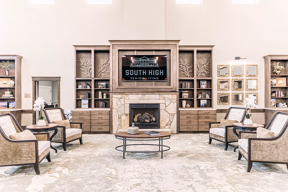 A bright and spacious senior living facility common area with a stone fireplace at the center, flanked by built-in wooden shelves and cabinets. Above the fireplace is a large screen displaying the logo and name 'South High Senior Living.' The room features four upholstered armchairs arranged around two round wooden side tables with white floral arrangements, and a round coffee table in the center. The walls are decorated with a set of square mirrors and decorative items on the shelves.