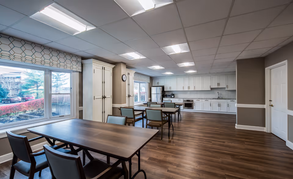 Communal dining room with wooden tables and chairs, large windows, and a long white kitchen along the back wall.