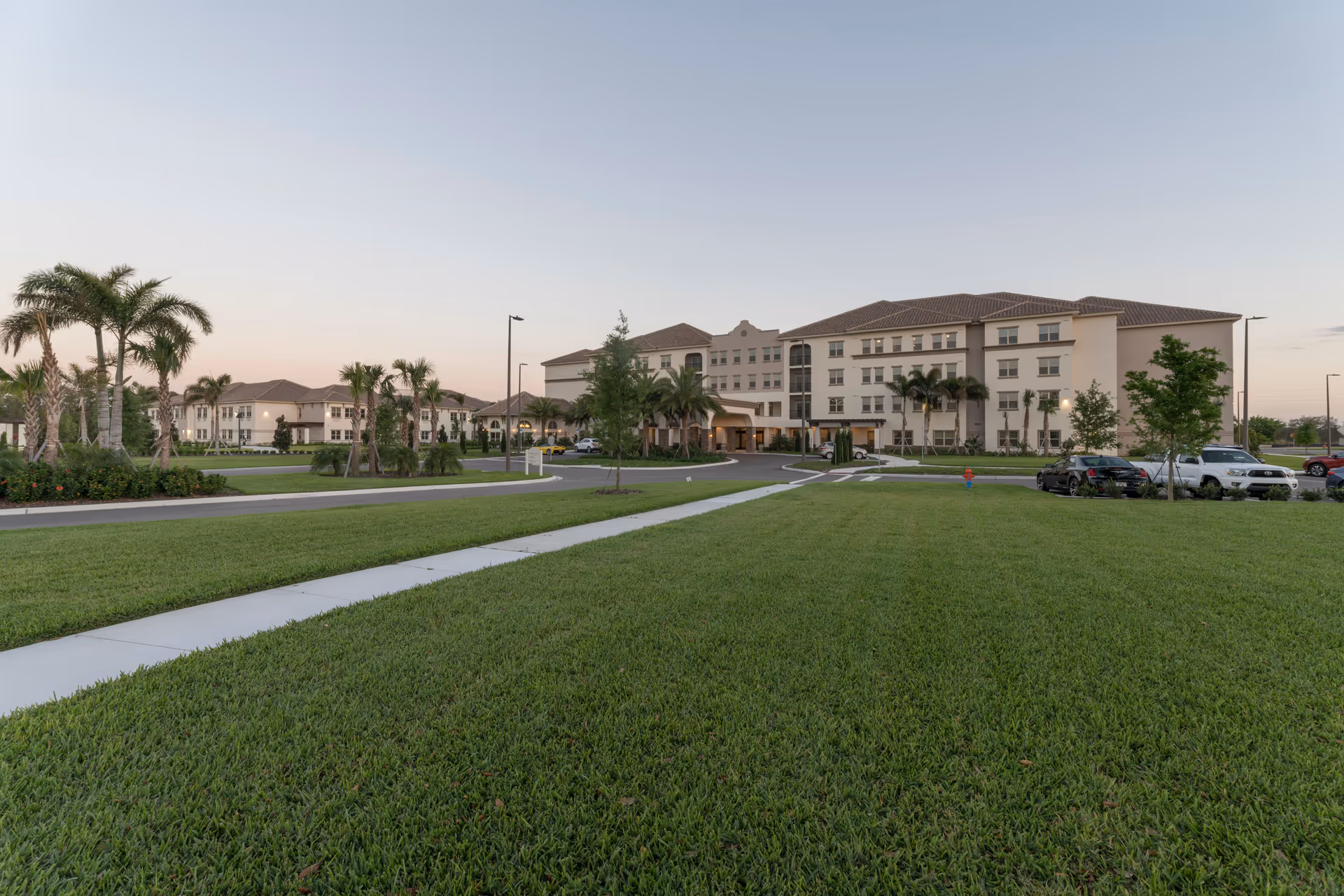 Front view of a large multi-story senior living building with a wide manicured lawn, palm trees, and a circular driveway.