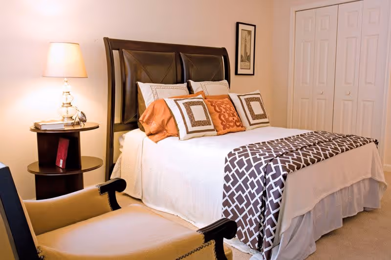 A cozy bedroom with a neatly made bed featuring a dark wooden headboard, white bedding, and decorative pillows in orange and brown patterns. A small round wooden side table with a lamp and books is beside the bed. There is a beige armchair in the foreground and a closet with white double doors in the background.