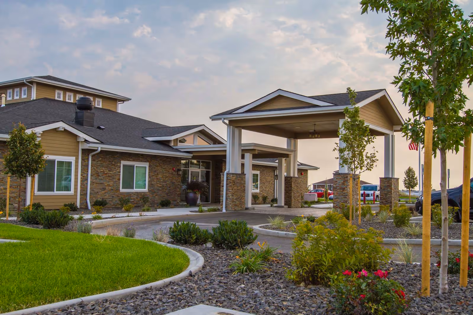 Exterior view of Fieldstone Memory Care Yakima facility showing a covered entrance with stone pillars, landscaped garden with small trees and bushes, and a driveway leading to the entrance under a partly cloudy sky.