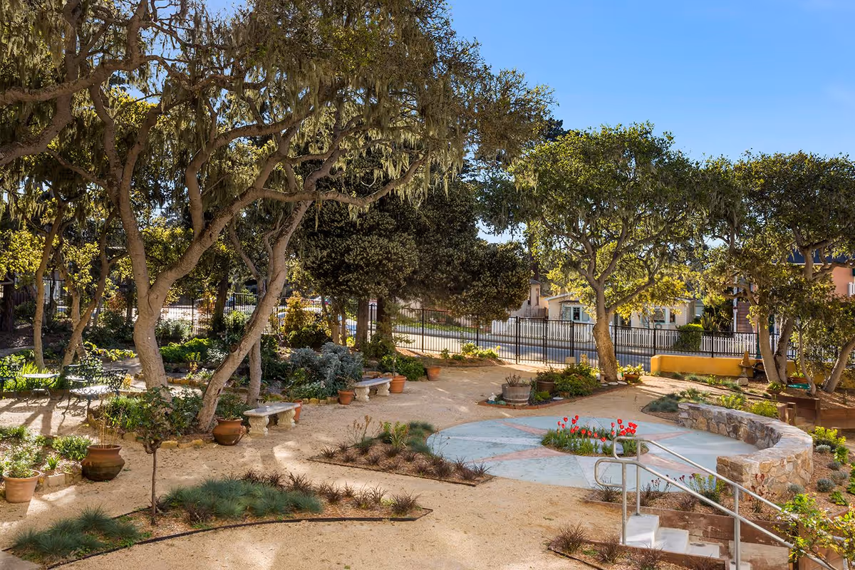 A sunny outdoor garden area at Pacific Grove Senior Living featuring mature trees with hanging moss, potted plants, benches, a circular paved area with flowers in the center, and a metal handrail along steps leading down to the garden path.