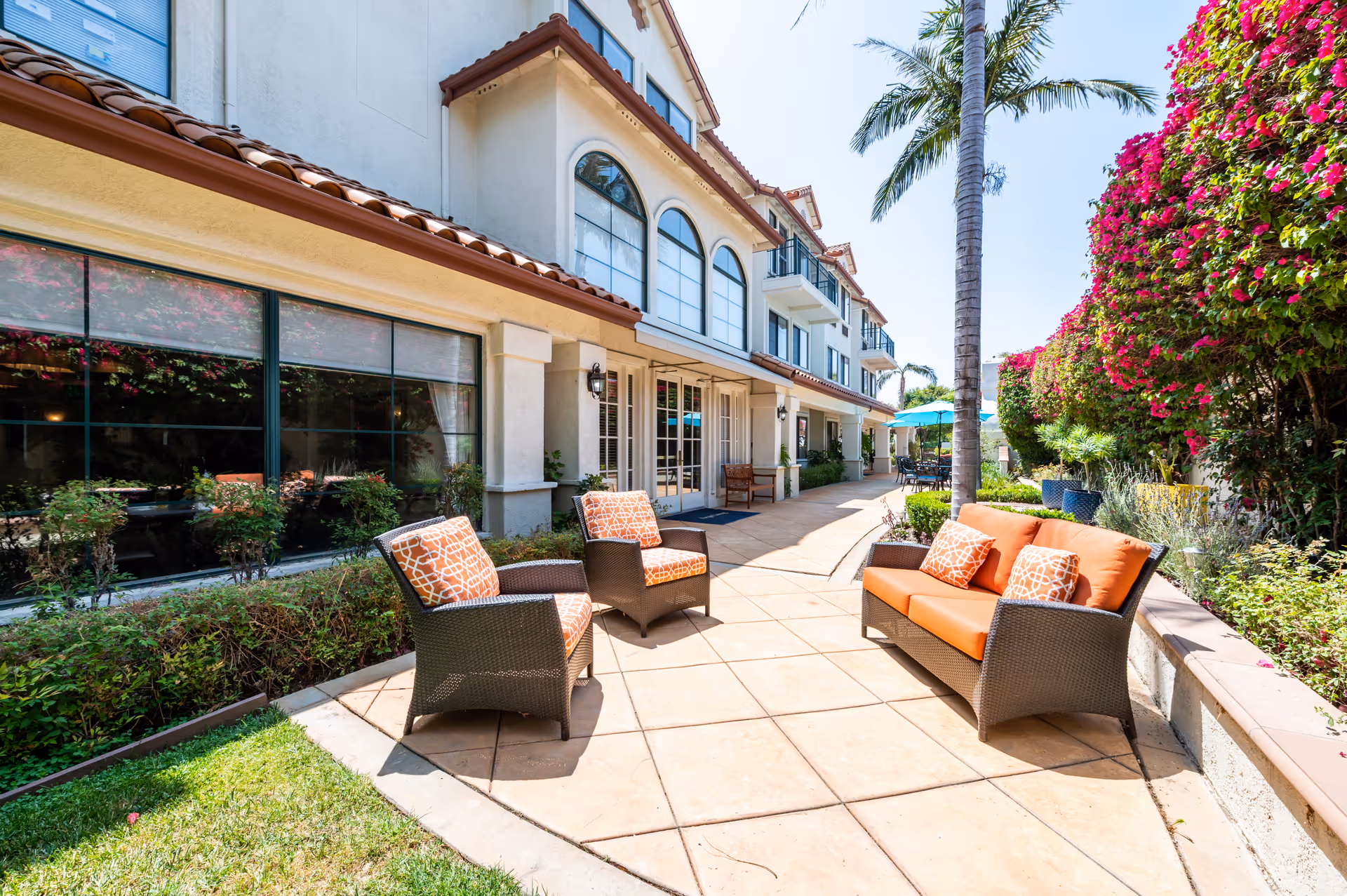 Sunlit outdoor patio with wicker chairs and a sofa with orange cushions beside a Mediterranean-style building, palm tree and flowering hedge.