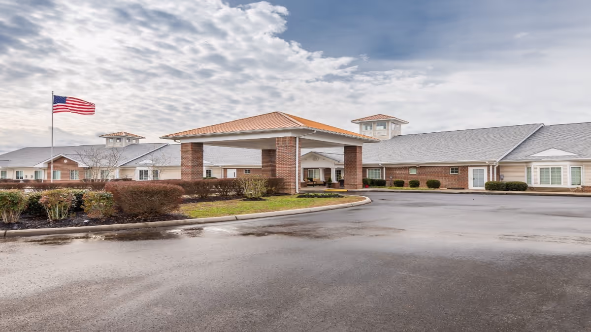 Exterior view of a senior living facility with a covered entrance supported by brick columns, a large American flag on a flagpole, and a paved driveway. The building has a combination of brick and siding with multiple windows and a gray shingled roof under a cloudy sky.