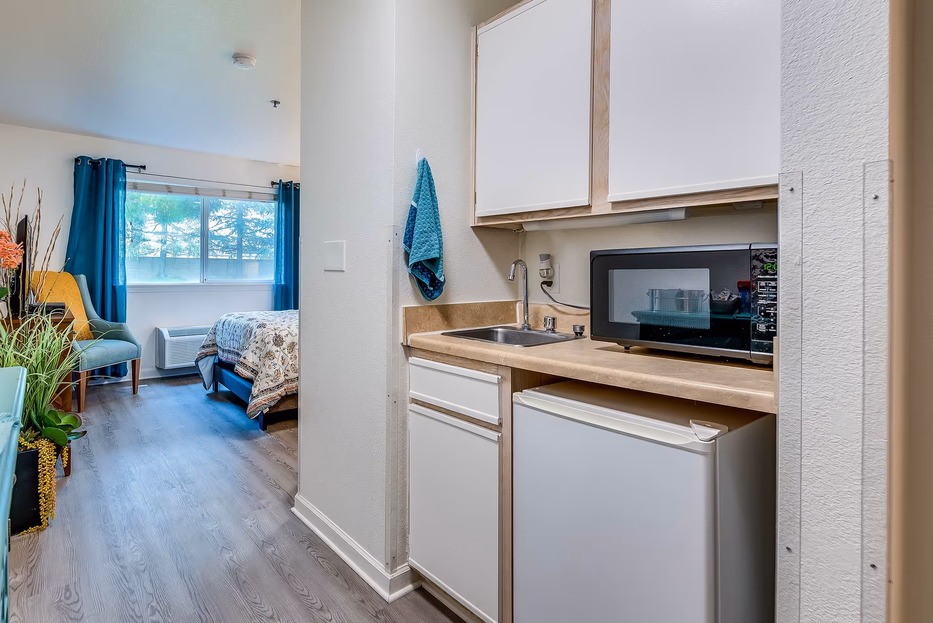 Small studio apartment interior showing a kitchenette with a microwave, sink and mini-fridge opening into a bedroom area with a bed, chair and window.