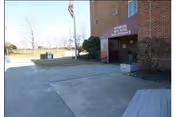 Exterior view of a brick building entrance with a maroon awning labeled 'Riverside Rest Home Main Entrance'. There is a concrete walkway leading to the entrance, a flagpole with an American flag, some bushes near the building, and an open outdoor area with trees and grass in the background.