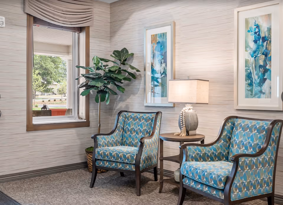 Two patterned armchairs flanking a side table with a lamp, a potted plant, framed artwork, and a window in a bright seating area.
