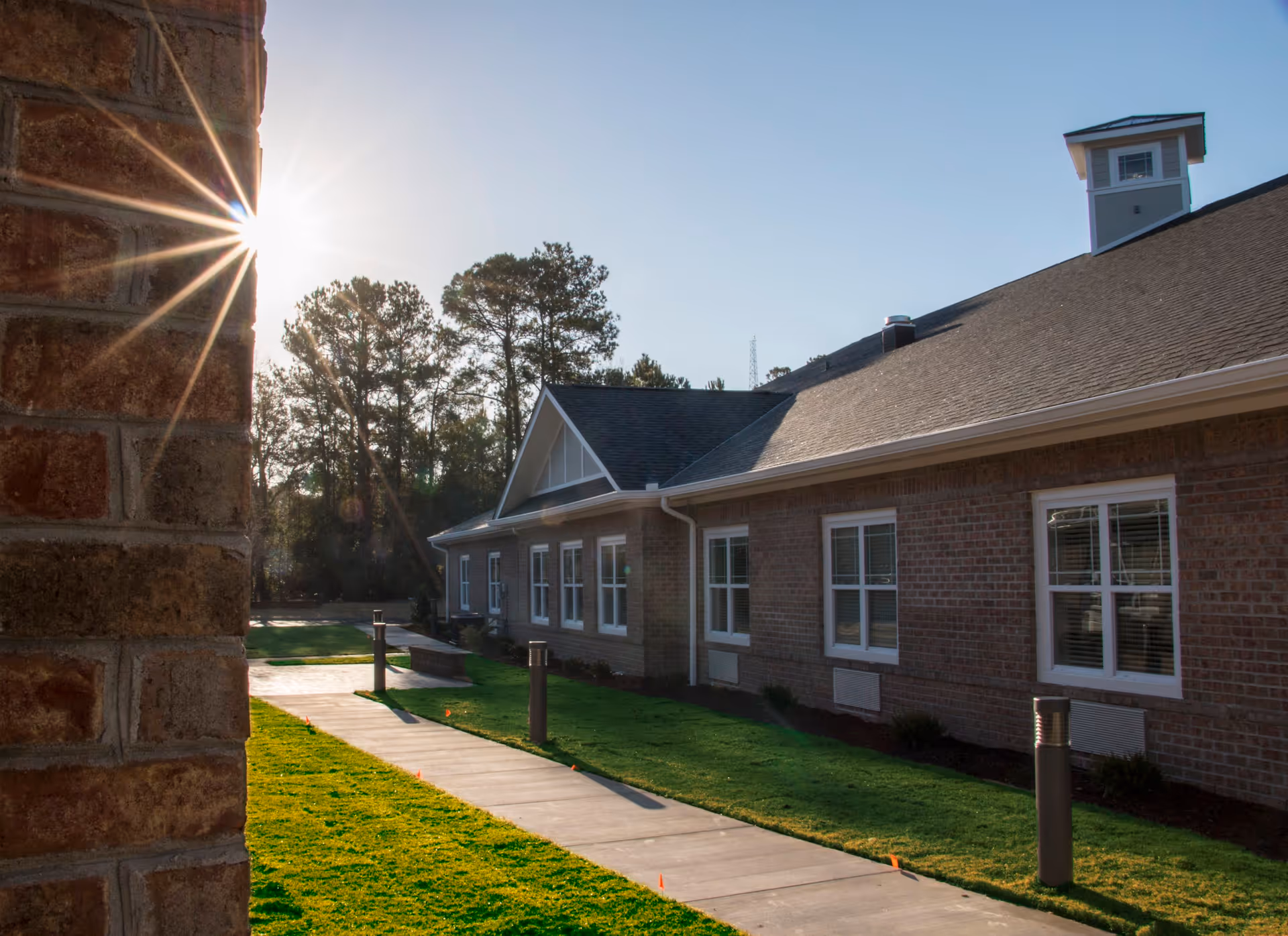 Exterior view of a brick building with multiple windows, a sloped roof, and a small tower-like structure on top. A concrete walkway runs alongside the building, bordered by green grass and small bushes. The sun is shining brightly from behind a brick wall on the left side, casting long shadows and creating a sunburst effect.