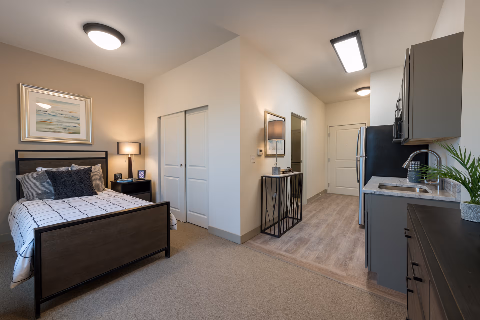 A modern senior living studio apartment featuring a bed with pillows and a nightstand with a lamp and clock. Adjacent to the sleeping area is a small kitchen with gray cabinets, a sink, a refrigerator, and a microwave. The flooring transitions from carpet in the bedroom area to wood in the kitchen and entryway. The walls are light-colored with framed artwork and a mirror above a small console table.