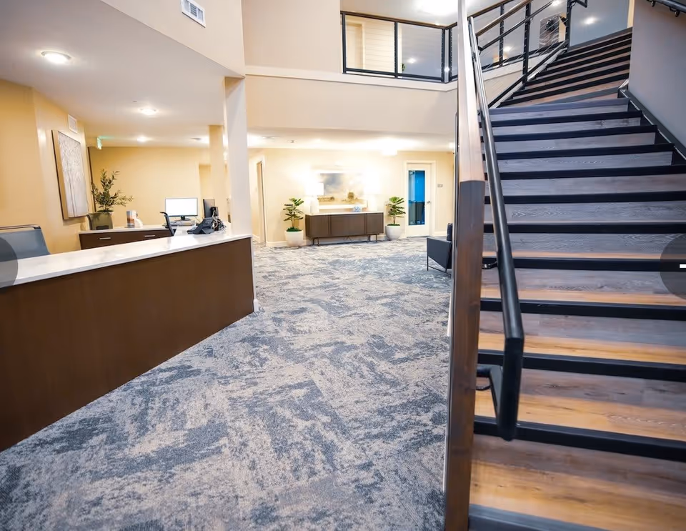 Interior view of a senior living facility lobby area with a reception desk on the left, carpeted floor, a staircase with wooden steps and black railing on the right, and a hallway with plants and furniture in the background.