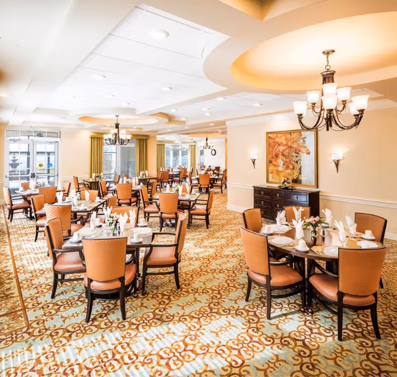 Spacious, well-lit dining room with multiple round tables set for meals, upholstered chairs, chandeliers, and patterned carpeting.