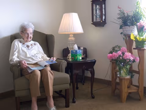 An elderly woman sitting in a cushioned armchair reading a magazine in a cozy room. Next to her is a wooden side table with a lamp, a box of tissues, and a phone. There are several flower arrangements on wooden stands near a window letting in natural light.