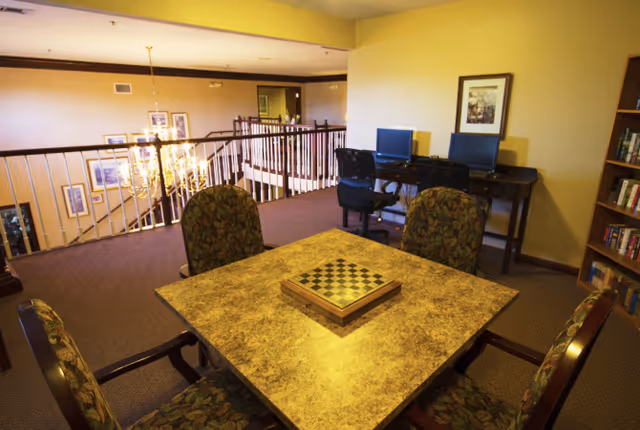 Interior common area with a square table and chessboard surrounded by upholstered chairs, computer stations against a wall, and a railing overlooking a lower-level chandelier.