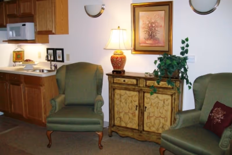 Interior view of a cozy living area in a senior living facility showing two green upholstered armchairs, a wooden cabinet with decorative carvings, a table lamp with a red and gold base, a framed floral painting on the wall, and a potted plant. To the left, there is a small kitchenette with wooden cabinets, a microwave, a sink, and some framed pictures on the wall.