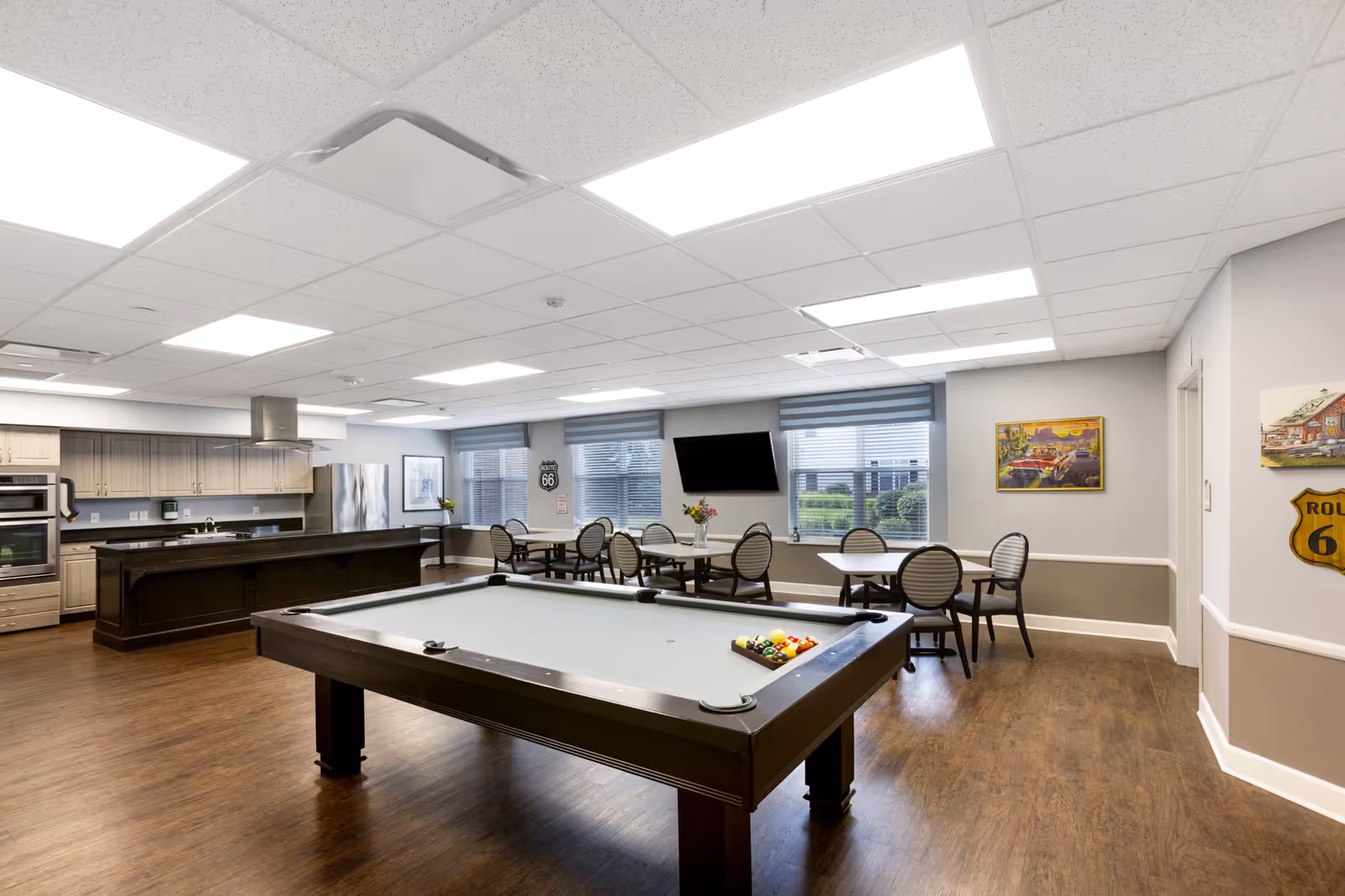 A spacious common area with a pool table in the foreground, several round tables with chairs, a wall-mounted TV, and a kitchen area with cabinets, a refrigerator, and an oven in the background. The room has large windows with blinds and Route 66 themed decorations on the walls.