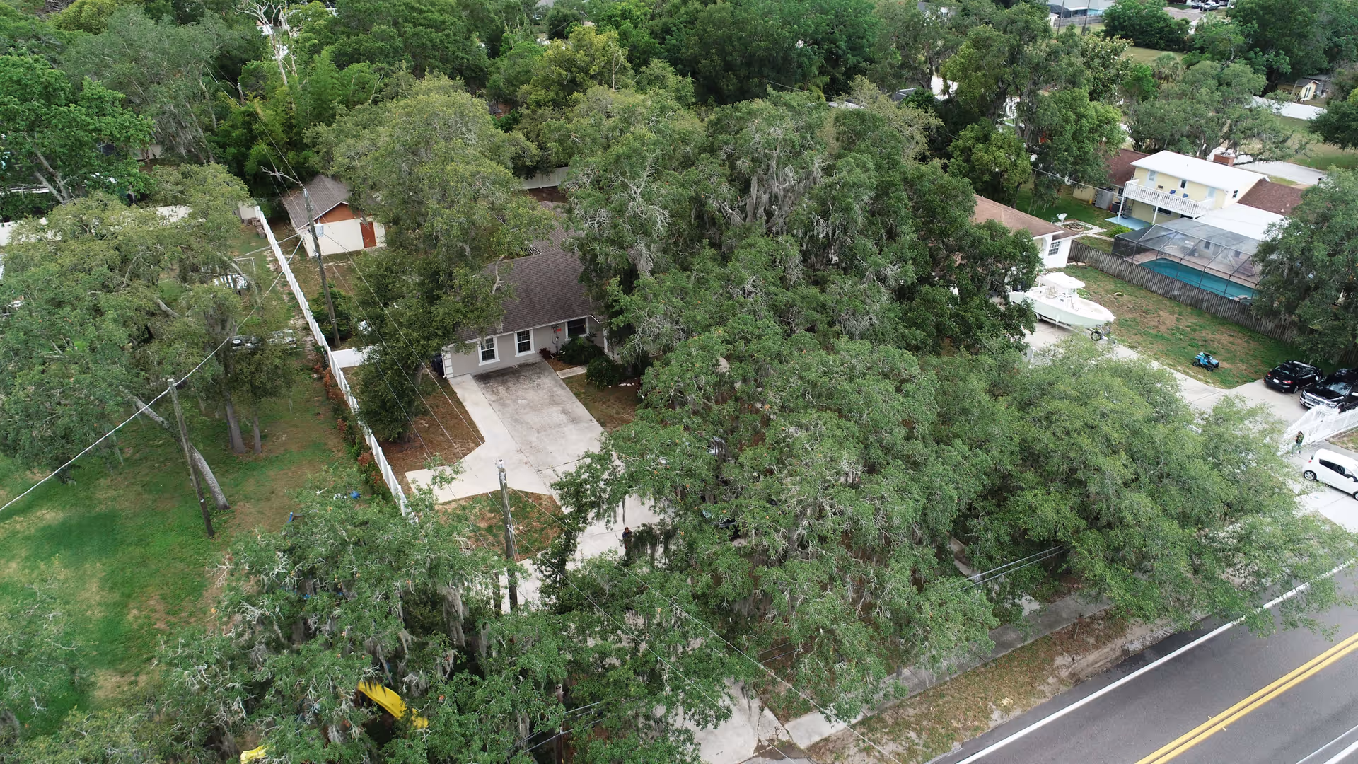 Aerial view of a residential area with a house surrounded by many large trees and greenery. The house has a driveway and is adjacent to a road with yellow dividing lines. Nearby properties include other houses, a boat, and parked cars.