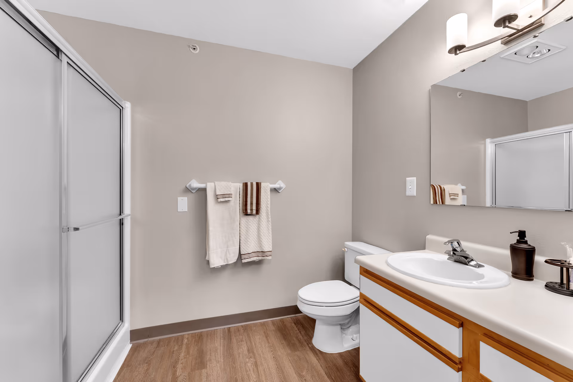 A clean bathroom with a shower enclosure with sliding doors on the left, a toilet in the center, and a vanity with a sink and mirror on the right. Two towels hang on a towel rack on the wall above the toilet. The floor has wood-like flooring and the walls are painted beige.