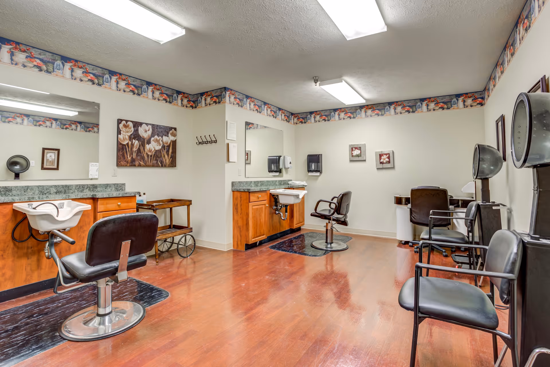 Interior view of a hair salon area in a senior living facility with wooden flooring, two hair washing stations with chairs, hair dryers, mirrors, and wall decorations including floral paintings and a decorative border near the ceiling.