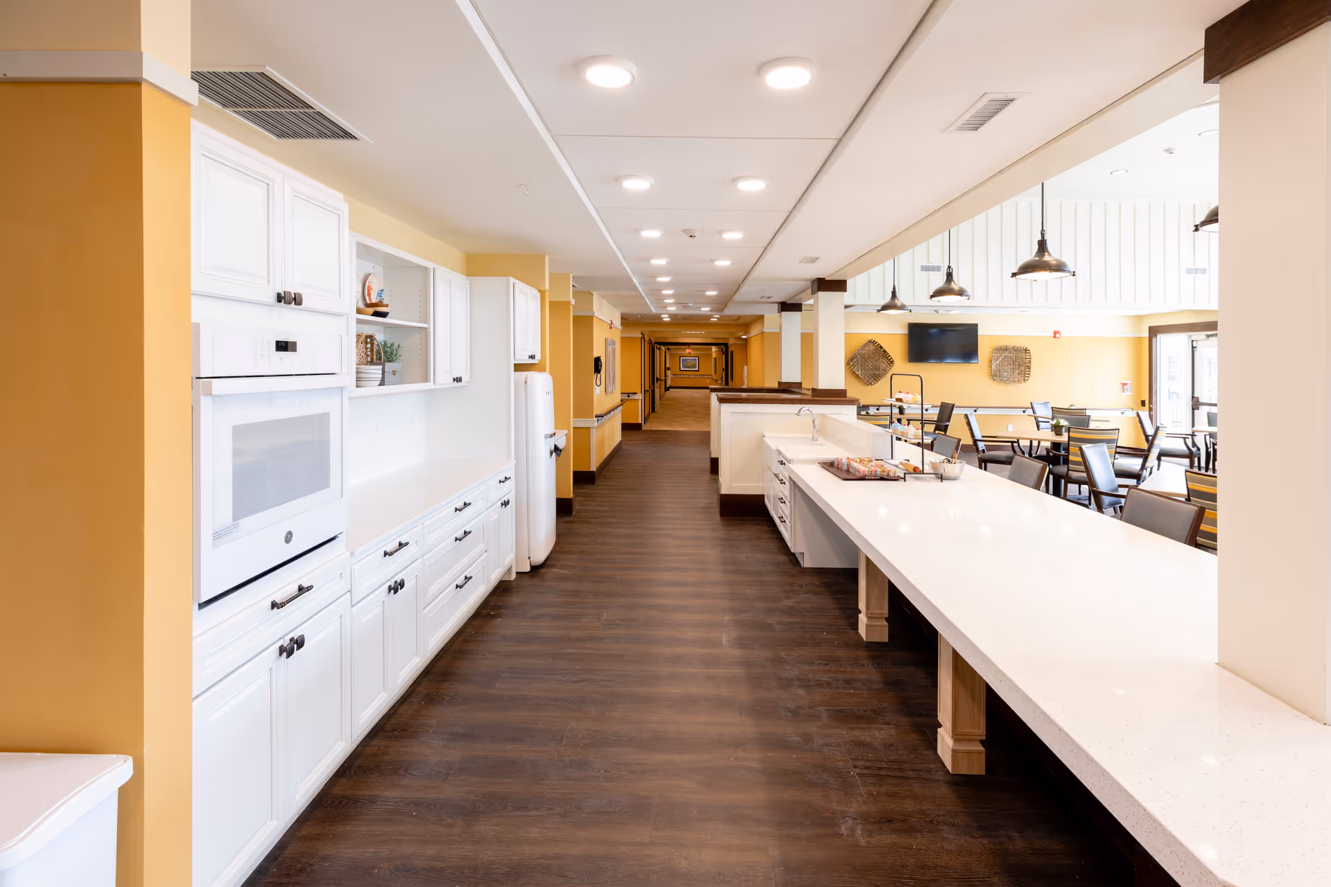 Bright open communal kitchen and dining area with a long white countertop, white cabinets, and seating in a senior living facility.