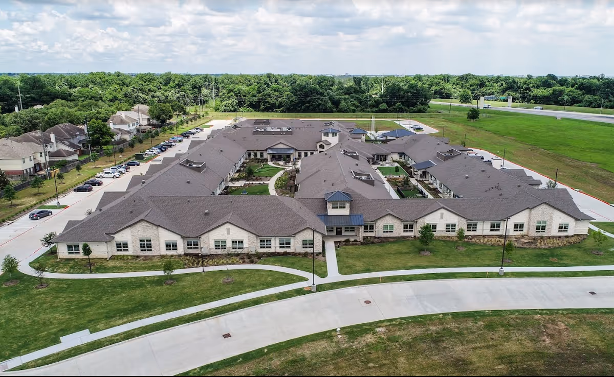 Aerial view of The Village at Sugar Land senior living facility showing a large, single-story building with multiple wings, surrounded by green lawns, trees, and parking areas. The sky is partly cloudy.