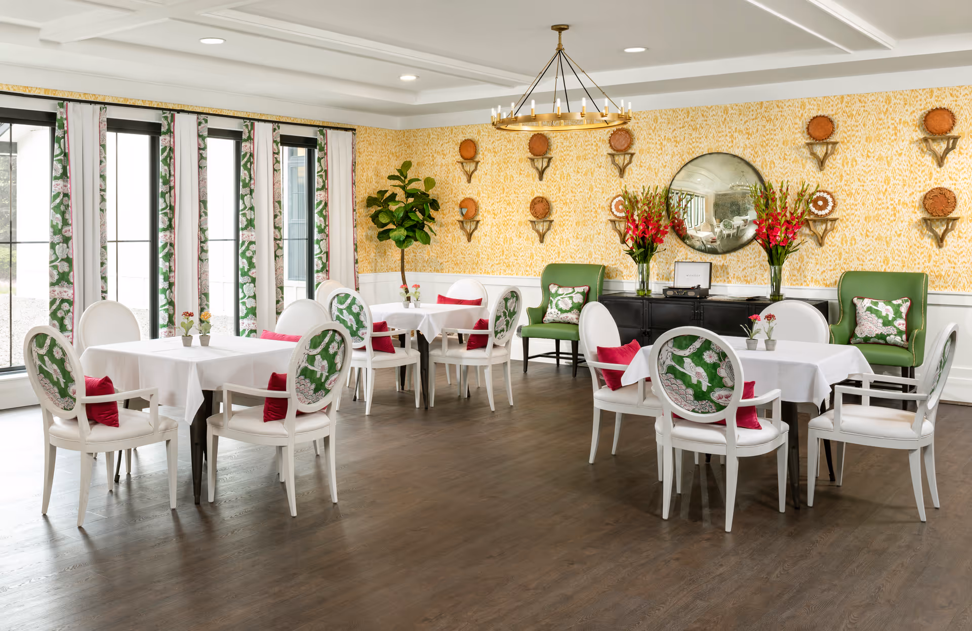 A bright and elegant dining room with several white tables covered with white tablecloths, surrounded by white chairs with green and pink patterned cushions. The room features large windows with floral curtains, a yellow patterned wallpaper, a round mirror on the wall, two green armchairs with decorative pillows, and floral arrangements on a black sideboard.