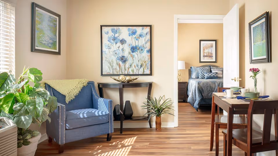 A cozy living area with a blue armchair draped with a light green throw blanket, a small wooden table with decorative items, and a potted plant. The room has light beige walls adorned with framed floral artwork. Through an open doorway, a bedroom with a neatly made bed featuring blue bedding and patterned pillows is visible. A small dining table with two chairs is set with dishes and a vase with a pink flower on the right side of the image. The floor is wooden with natural light streaming in from a window on the left.