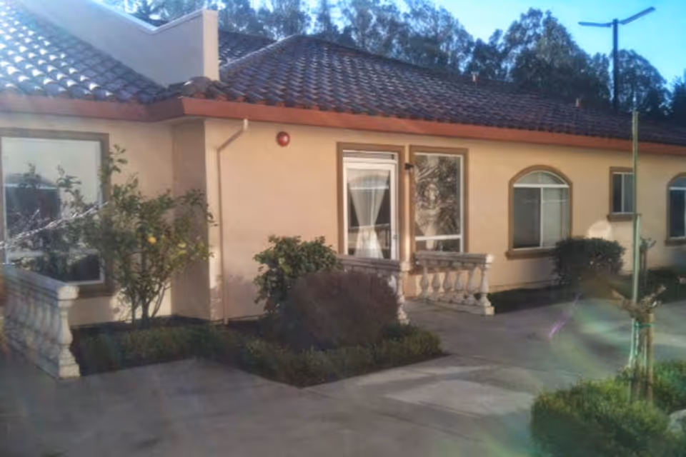 Exterior view of a single-story building with a tiled roof, beige walls, and several windows. There are small bushes and plants along the building, and a concrete pathway in front. The scene is outdoors with trees visible in the background under a clear sky.