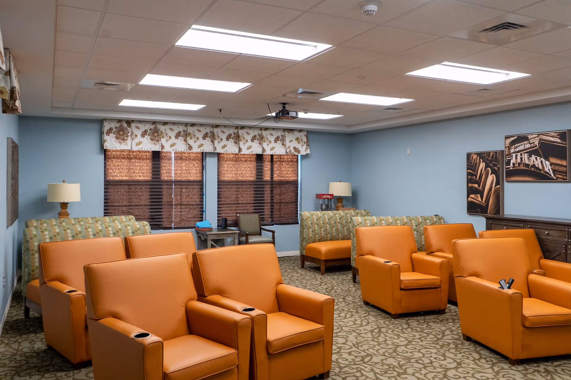 A cozy common room with multiple orange leather armchairs arranged in rows facing a projector. The room has light blue walls, patterned carpet, two windows with floral valances and brown blinds, and framed pictures on the wall. There are also two patterned sofas and two table lamps in the background.