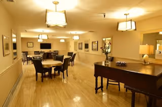 A well-lit common area in a senior living facility with multiple round tables and chairs arranged for seating. The room features warm lighting with ceiling lamps, framed pictures on the walls, a piano with a vase of flowers on top, and a television mounted on the far wall.