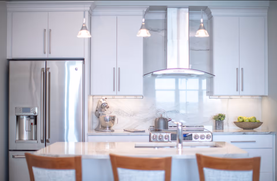 Modern kitchen with white cabinets, stainless steel refrigerator, stove with a kettle, a stand mixer on the counter, a bowl of green apples, and three wooden chairs at a marble island with a sink and faucet.